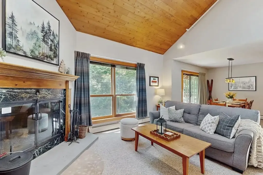 Living room seating area facing a large window, with a stone-look fireplace and vaulted wood ceiling